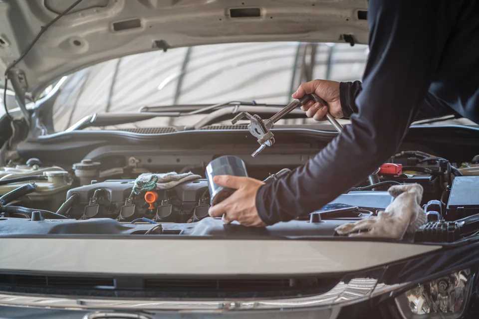 Mechanic working under an open bonnet in a well-lit workshop bay, using a hand tool on the engine.